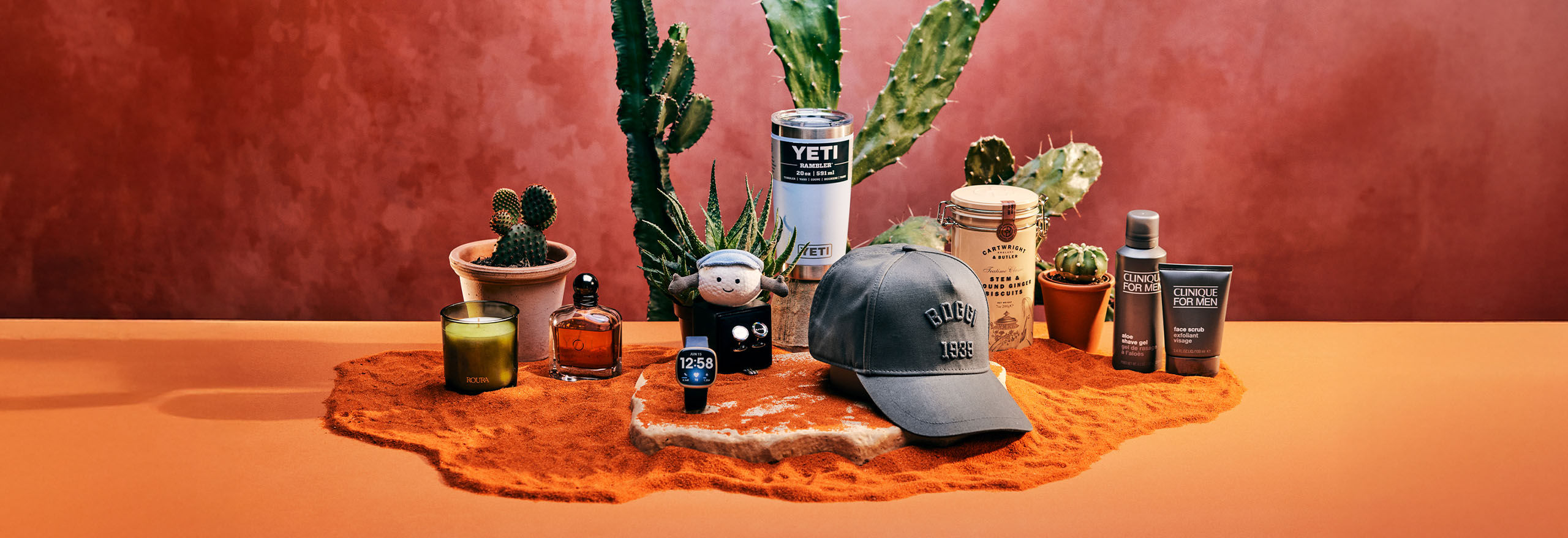 A hat, skincare products, a candle, and cacti are arranged on a table with a reddish-brown background.