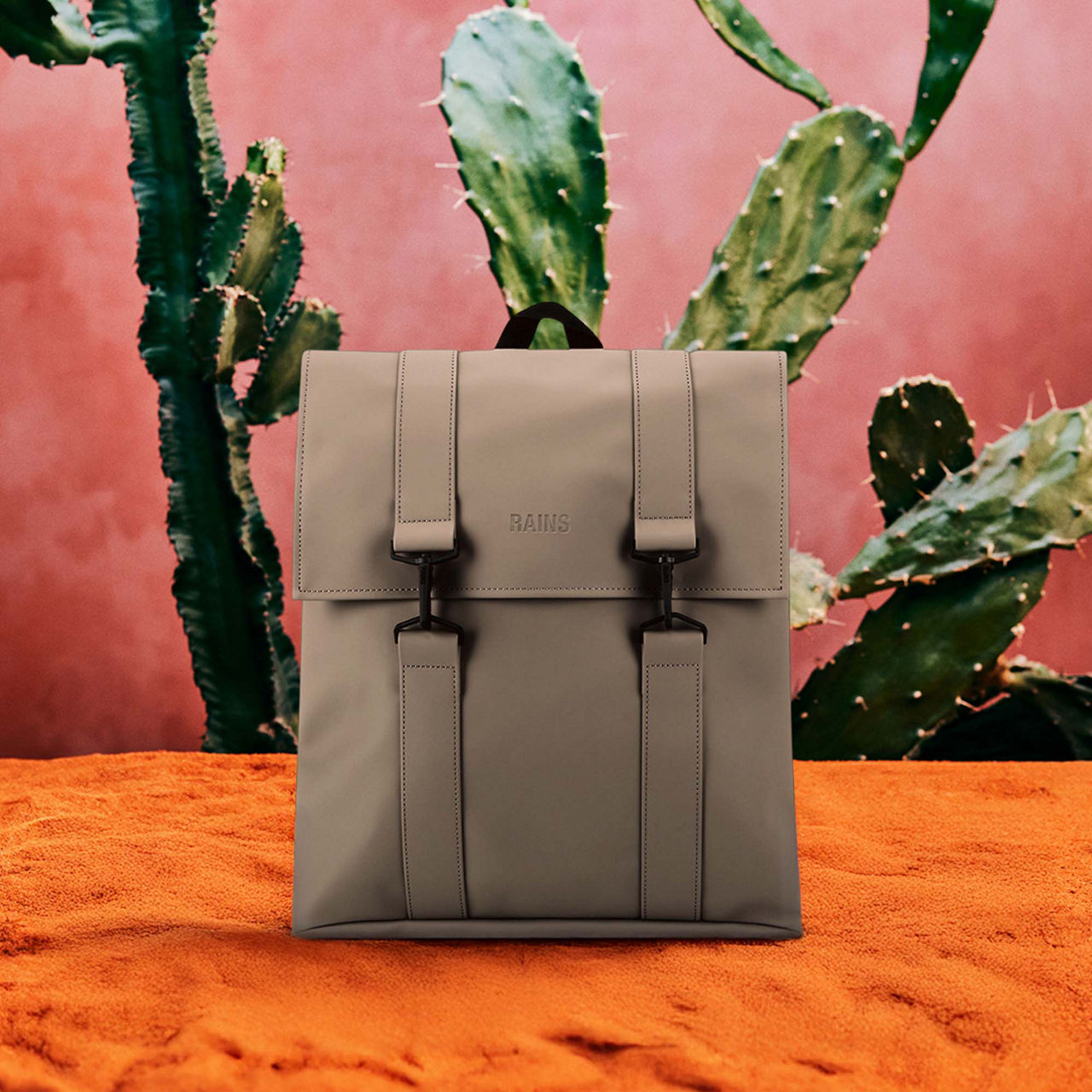 A gray backpack is displayed against a background of green cacti on a pink wall.