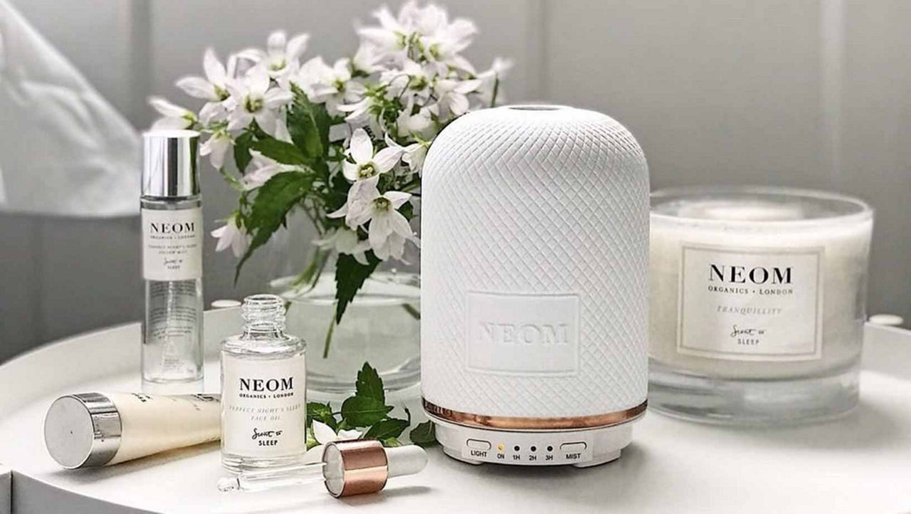 NEOM products including a diffuser and a candle on a white side table with white flowers in the background.