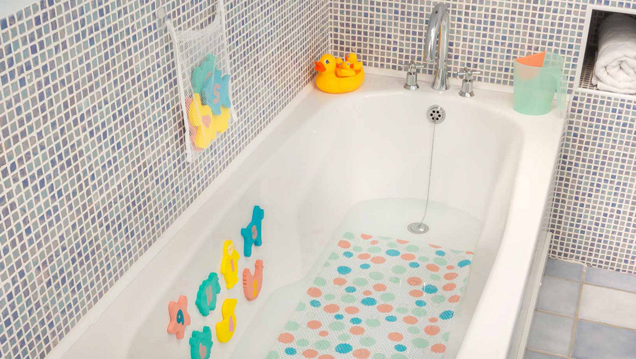 A white bath with baby bath toys sitting on the edge and hanging from a white mesh bag on the blue tiled wall. The toys include rubber ducks and colourful animals with numbers on them. These animals can also be seen suck to the inside of the bath.