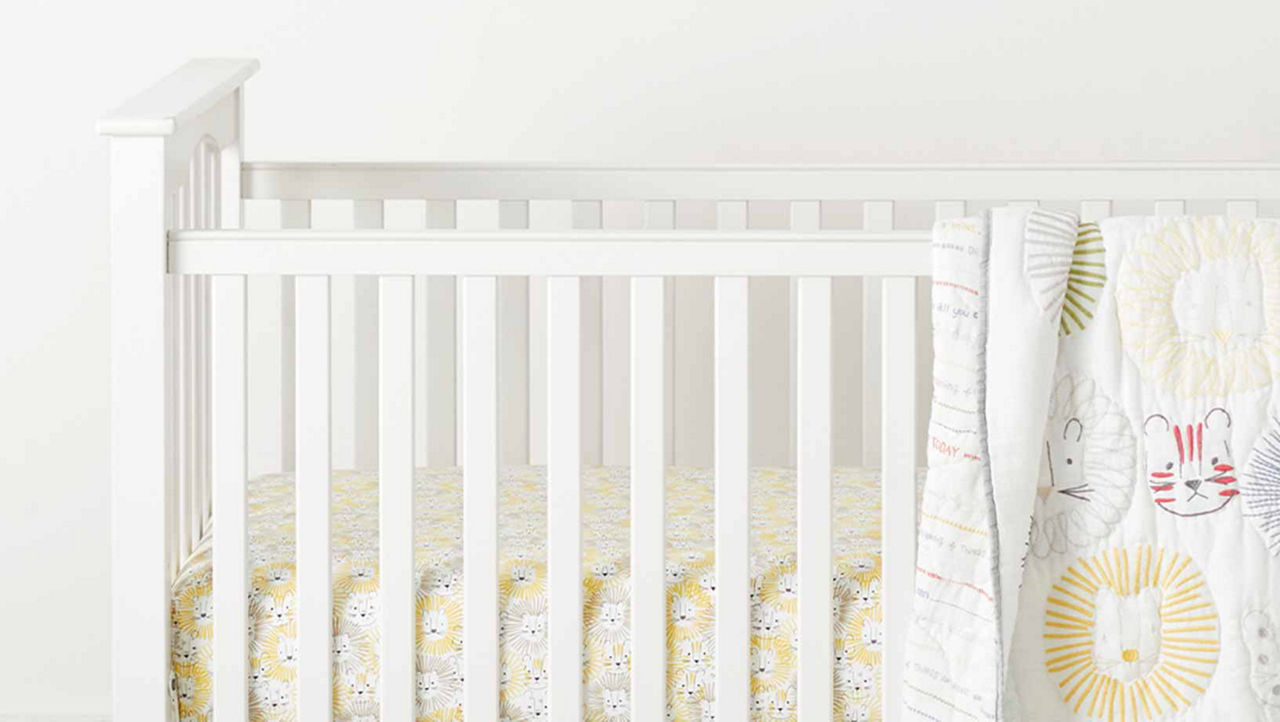 A white cot with a white baby blanket featuring embrodiered lions and tigers in yellow ann red hangs over the side. The mat inside the cot is covered in a sheet that matches the pattern of the blanket but is all in yellow.