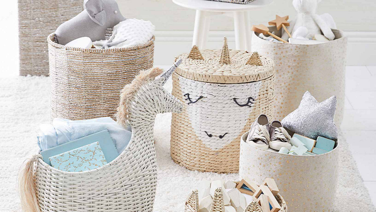 Six wicker baskets are sitting on a white furry rug. Two of the baskets have the design and shape of a unicorn on them. All the baskets are filled wth blue and grey blankets and soft toys.