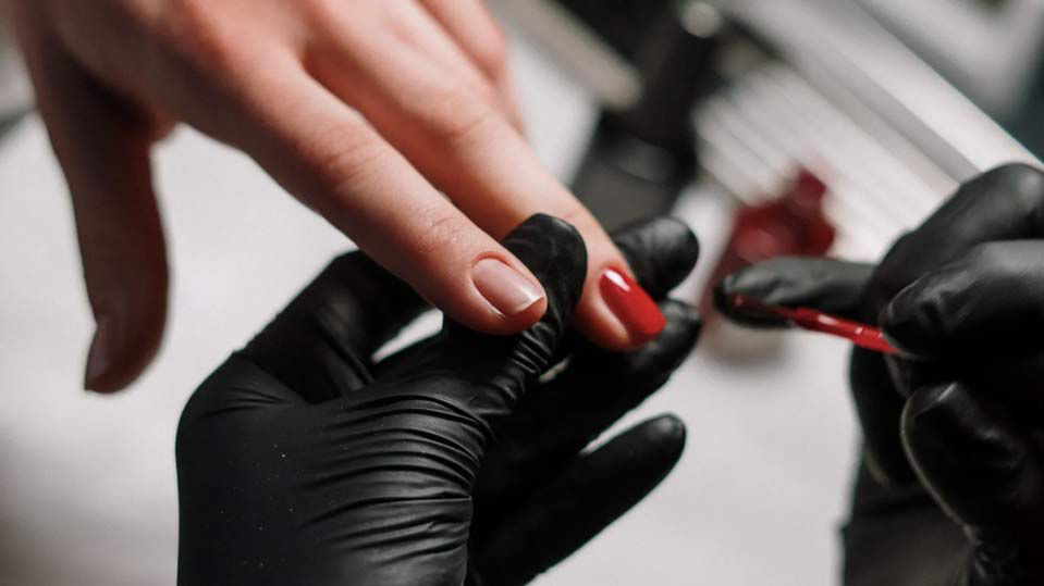 close up of a nail technician wearing gloves painting a woman's nails in red nail polish
