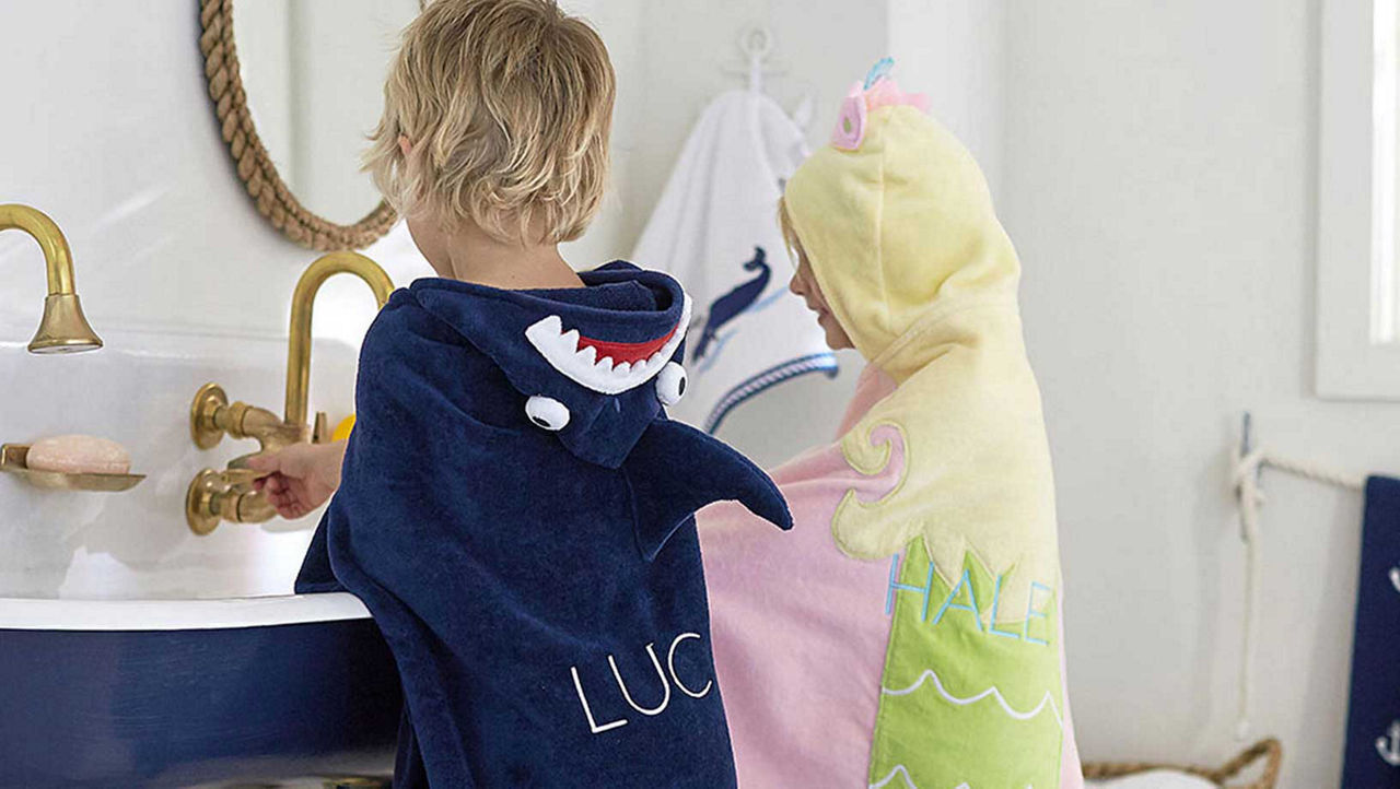 A young boy and girl wearing Pottery Barn Hooded Towels while washing their hands at a navy and white skink. The boy wears a navy towel with a skark on the hood and the girl wears a pink yellow blue and green towel with a unicorn on the hood.